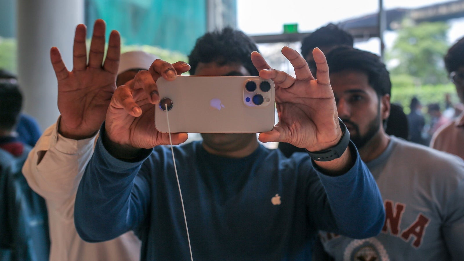 A group of people is seen in a store, with one person in the foreground holding up a smartphone for a photo. The individual is wearing a blue long-sleeve shirt featuring an Apple logo, while others can be seen behind him.
