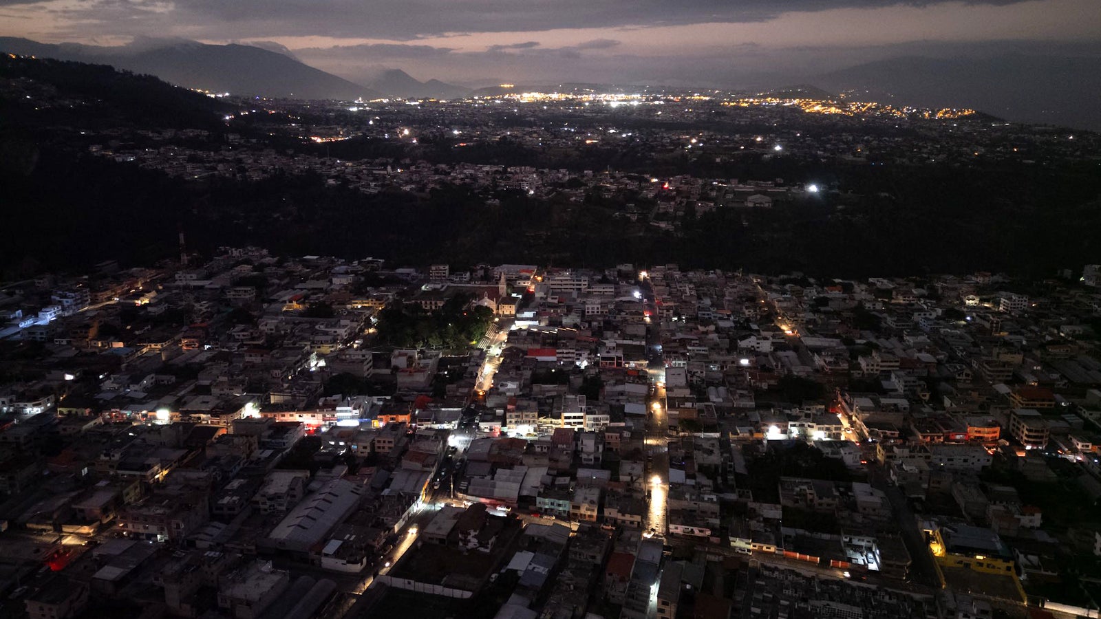 Aerial view of a city at night, with a mix of well-lit and dark areas, showcasing dense housing, streetlights, and distant city lights against a backdrop of mountains under a cloudy sky.