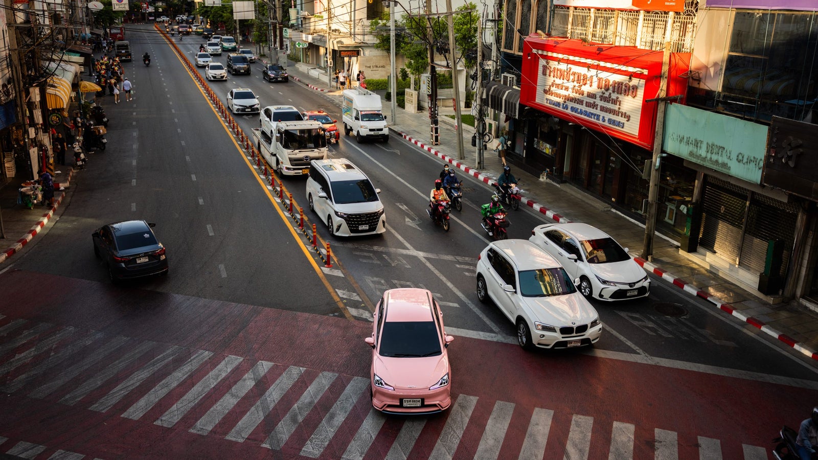 A busy urban intersection with vehicles including cars, motorcycles, and vans moving in various directions, featuring a prominent pink car in the foreground on a crosswalk, amidst hustle and bustle of city life.