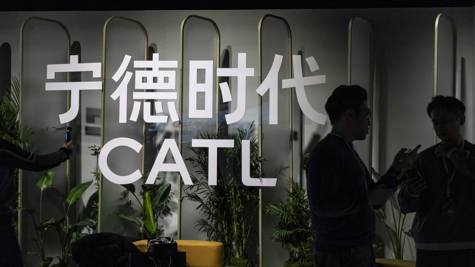 A modern display featuring large white lettering in Chinese and the abbreviation 'CATL' on a dimly lit backdrop, with greenery and furniture visible in the foreground. Two silhouetted individuals are engaged in conversation near the display.