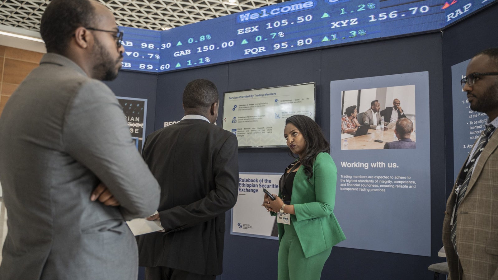 A woman in a green suit discusses with two men in business attire at a booth displaying information about the Ethiopian Securities Exchange, featuring a digital screen with stock prices and details about services offered to trading members.