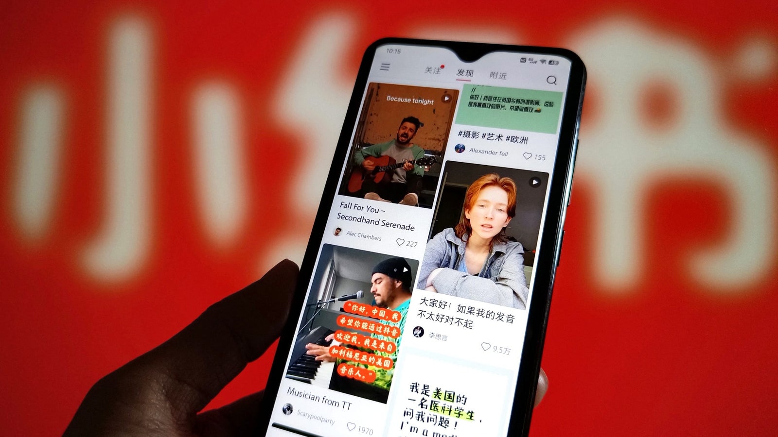 A close-up of a smartphone held in a hand, displaying the social media app interface of Xiaohongshu with videos and user profiles. The background is a blurred red with white text.