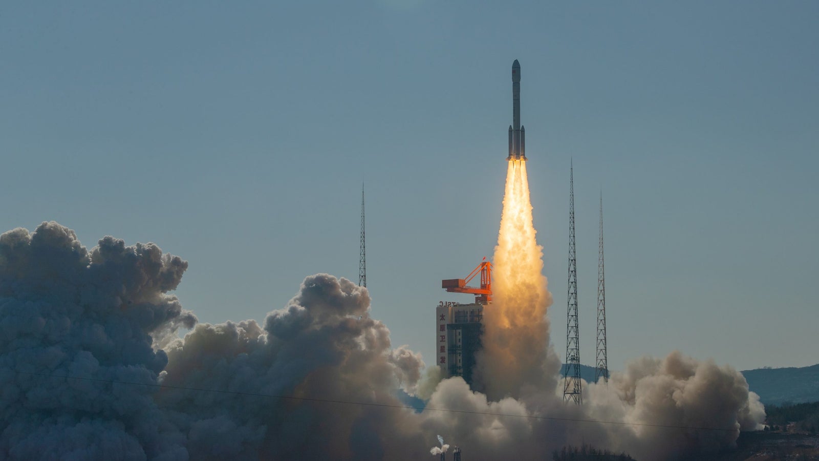 A rocket launches into the sky, leaving behind a plume of smoke and flames, with tall communication towers visible in the background under a clear blue sky.