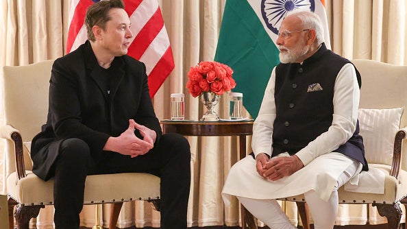 Elon Musk and Indian Prime Minister Modi seated face-to-face in a meeting, surrounded by flags of the USA and India.