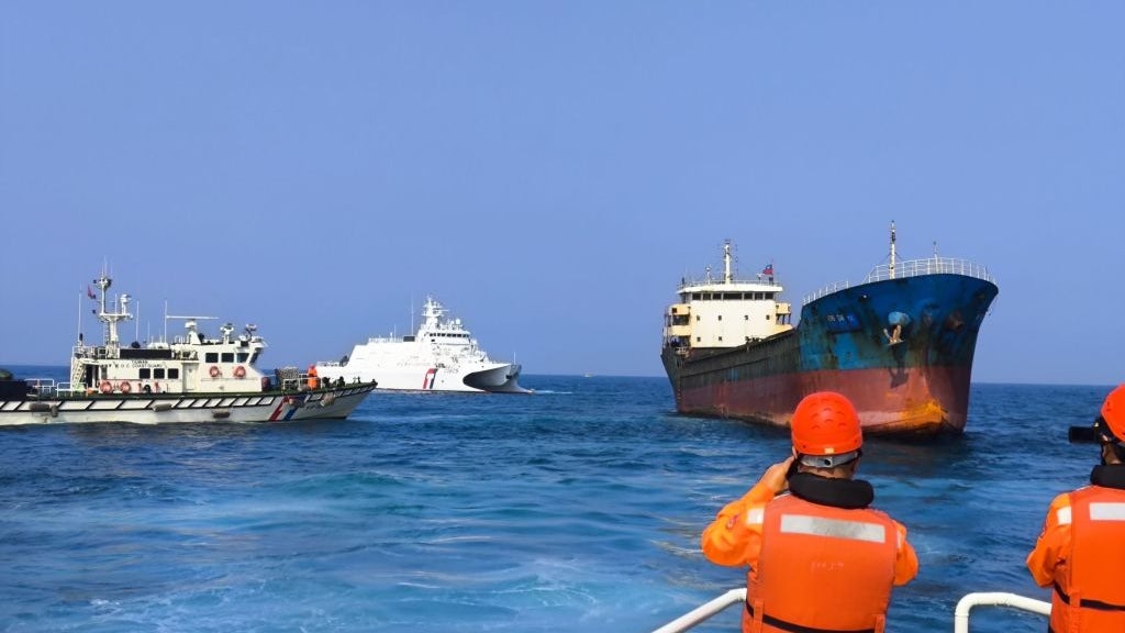 Two patrol boats and a larger cargo ship in open water, with a clear blue sky overhead; two people in orange safety vests stand on the deck of one vessel, observing the scene.