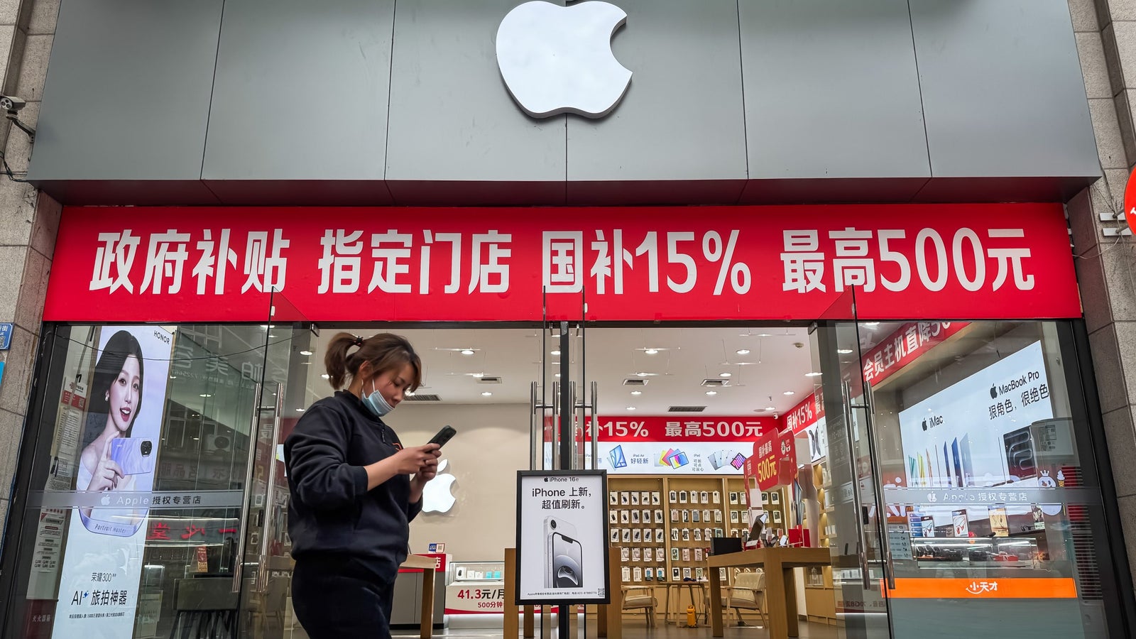 An Apple store entrance featuring large red promotional signs highlighting discounts, with a young woman in a black hoodie and mask looking at her phone while walking by.