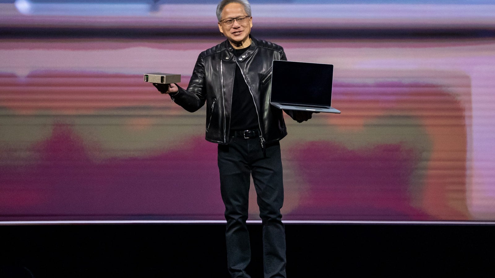 A man in a black leather jacket stands on stage holding a compact device in one hand and a laptop in the other, with a colorful digital backdrop behind him.