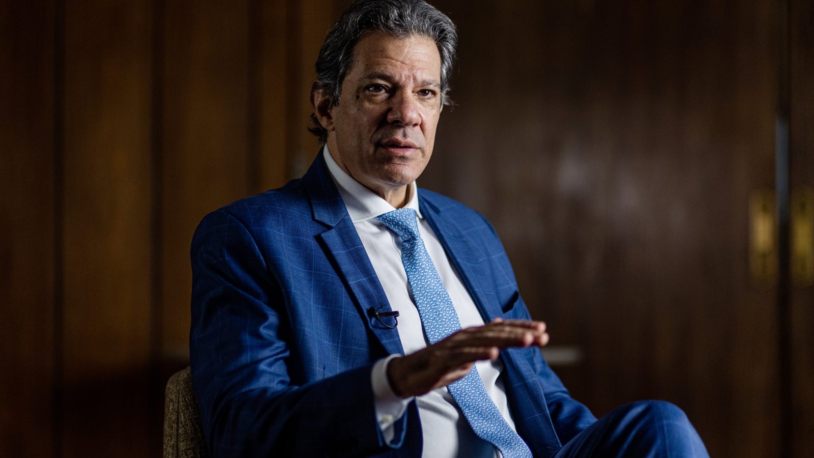 A man in a blue plaid suit and a light blue tie gestures with his right hand while sitting in a chair, conveying a serious expression against a wooden backdrop.