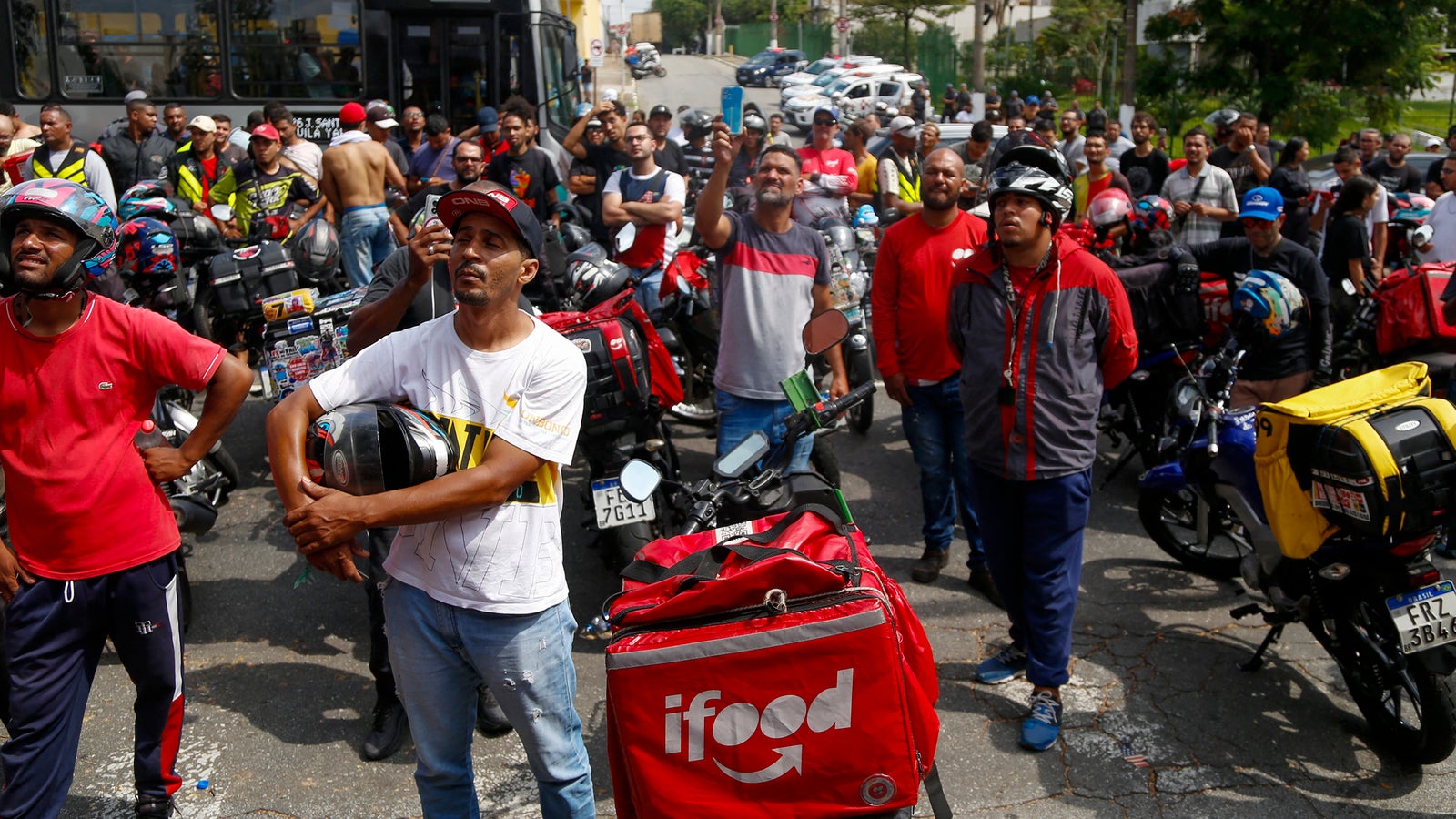 A group of delivery riders, wearing helmets and colorful uniforms, gather in a street, some holding helmets and others looking towards the sky, while motorcycles with delivery bags marked 'ifood' are lined up in the background.