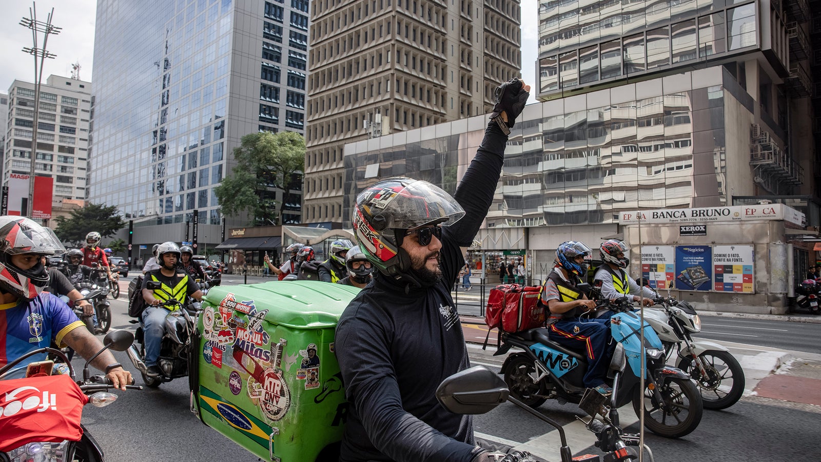 A group of delivery motorcyclists in a busy urban setting, with one rider wearing dark sunglasses and raising his hand. The motorcycle beside him has a green delivery box covered in stickers, while other riders in bright vests are also present. Skyscrapers reflect in the background.