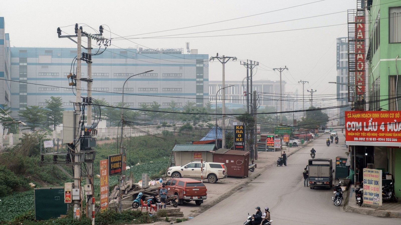 A street scene in an industrial area with power lines, several parked vehicles, and a restaurant sign visible. The background features a large, modern building with a gray sky, indicating a hazy atmosphere.