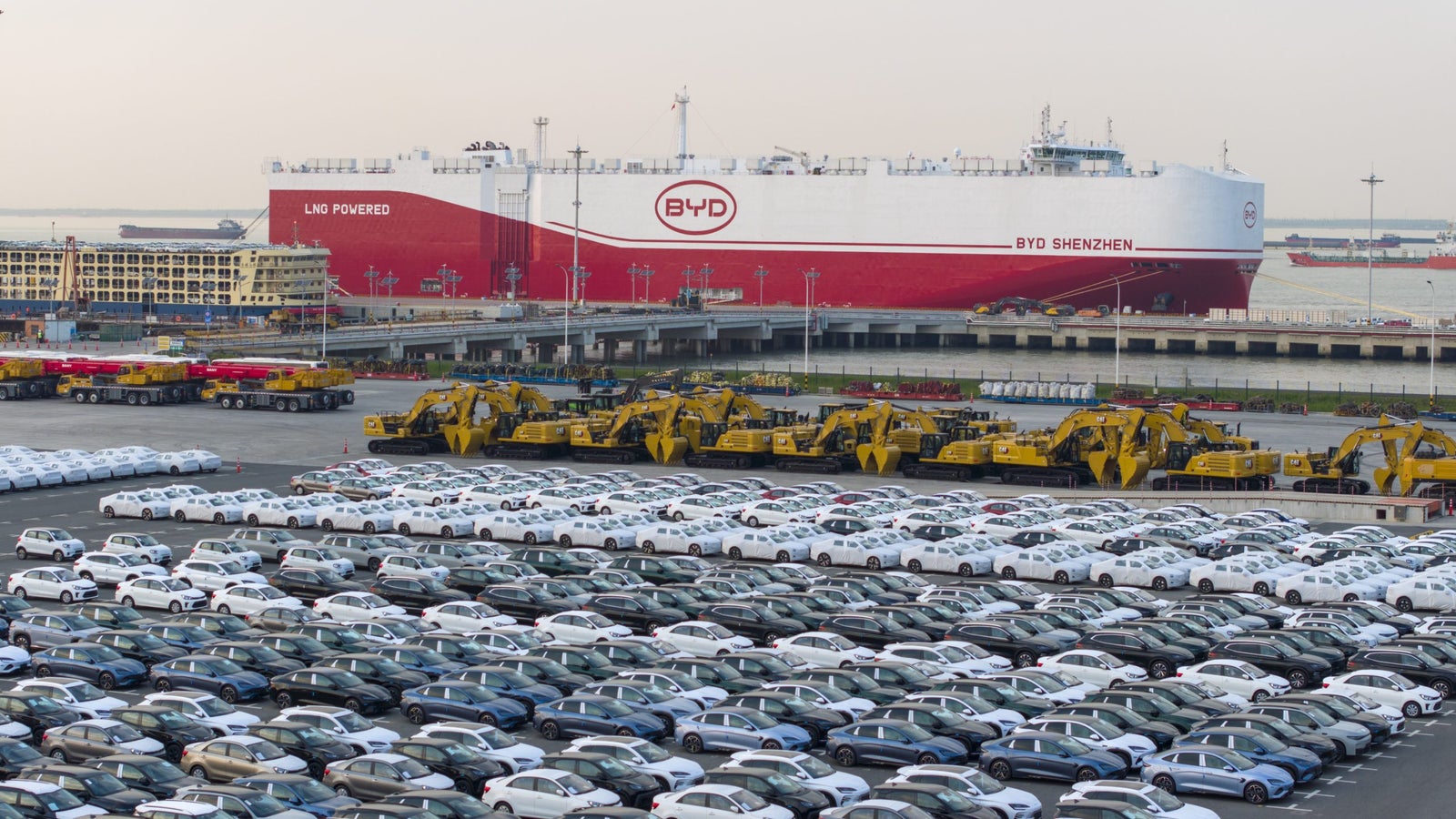 A busy port scene featuring rows of parked cars in various colors, with a large cargo ship displaying the BYD logo docked in the background. Yellow heavy machinery is also visible near the vehicles.