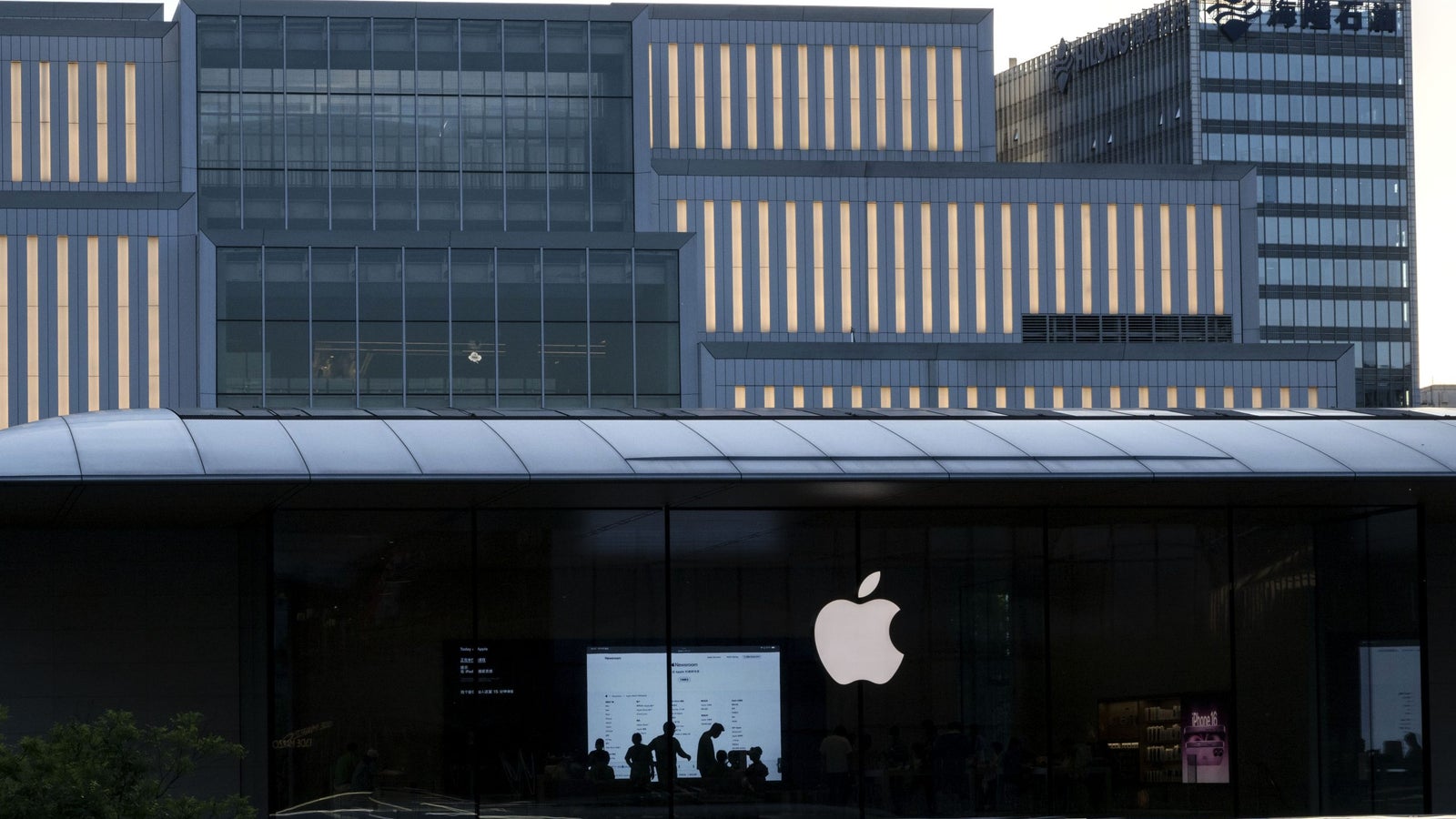 The exterior of a modern Apple Store featured prominently with a large Apple logo, showcasing a glass front reflecting the outlines of a contemporary building, illuminated windows, and silhouettes of people visible inside the store.