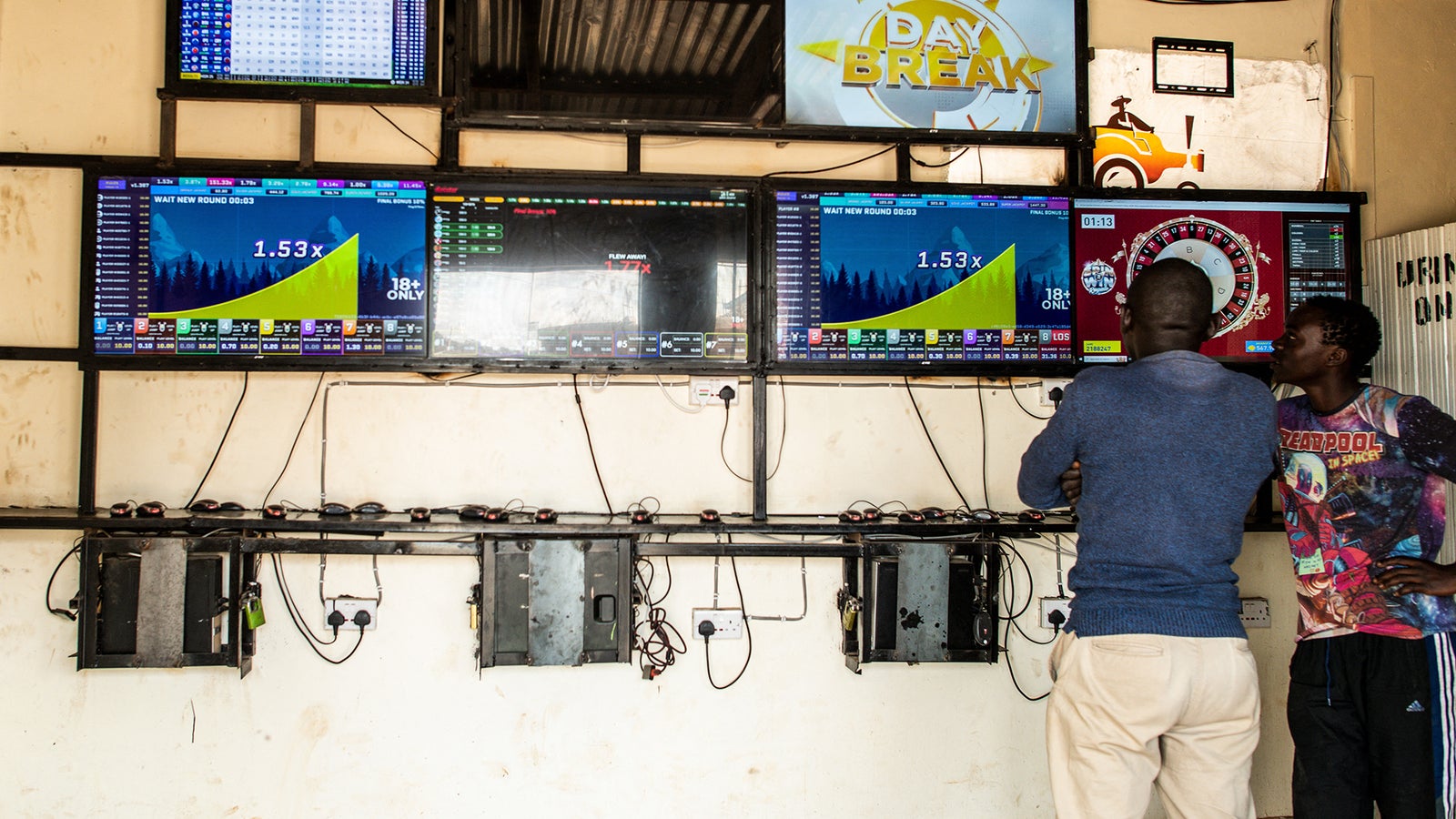 Two men are standing in front of multiple large screens displaying betting information and statistics in a dimly lit room.