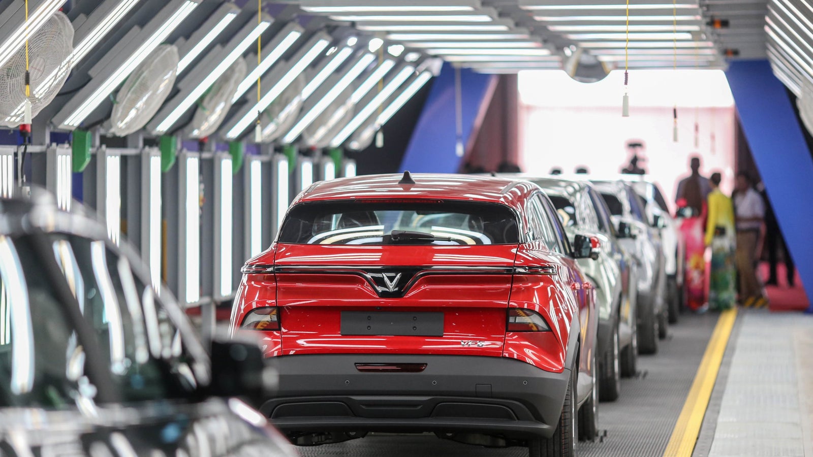 A red SUV parked in a modern car production facility, with multiple vehicles lined up in the background, illuminated by bright fluorescent lights.
