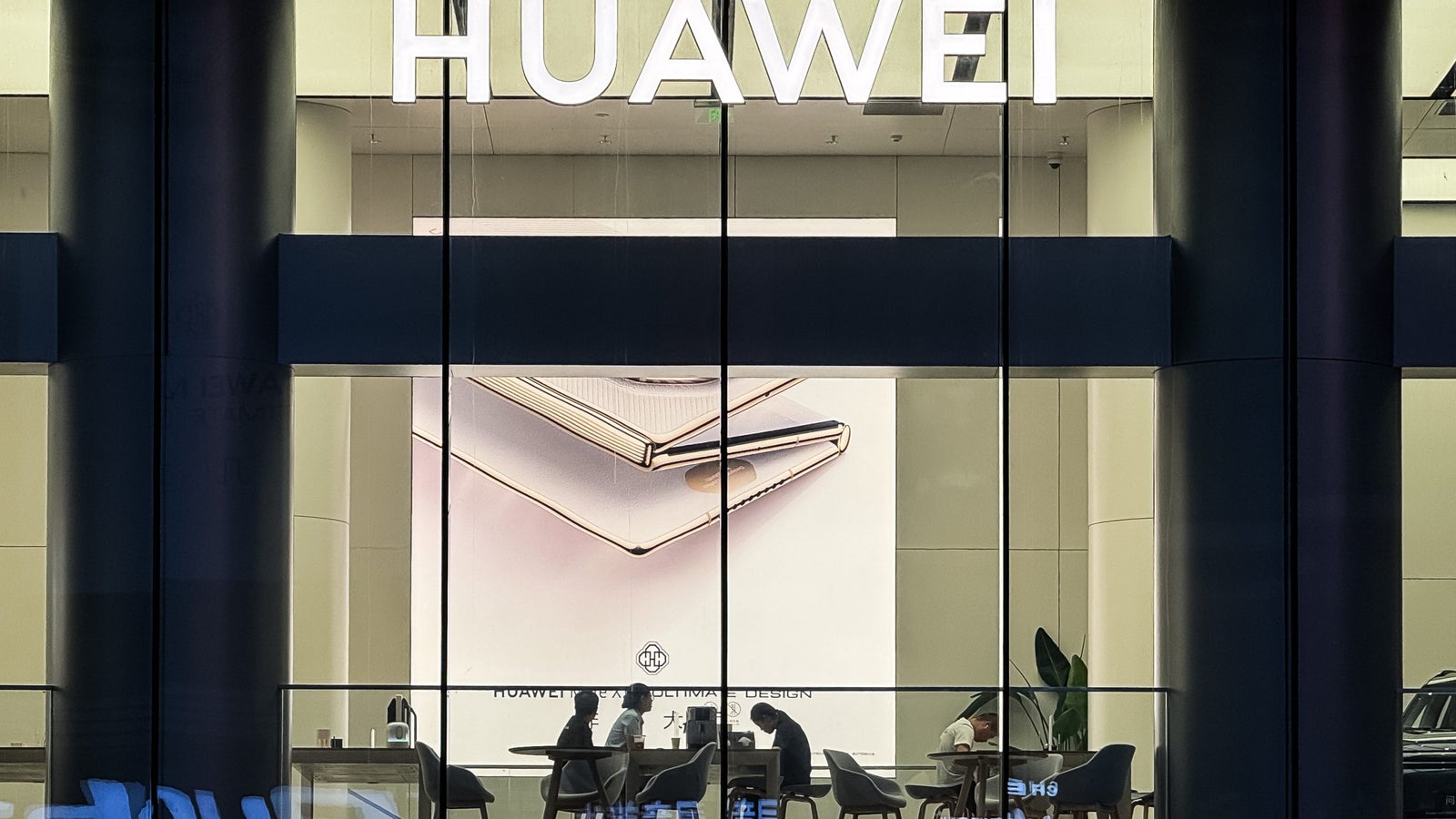 View of a Huawei store featuring a large advertisement of a smartphone on the wall. Inside, several people are seated around a table, engaged in conversation.