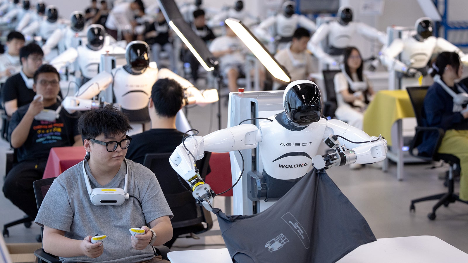 A robot assists a man as he folds a shirt, surrounded by other robots and people in a modern workspace.