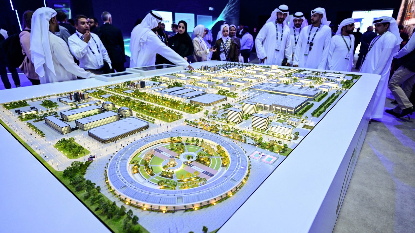 A detailed model of a futuristic city layout, with people observing and discussing around it at an exhibition.
