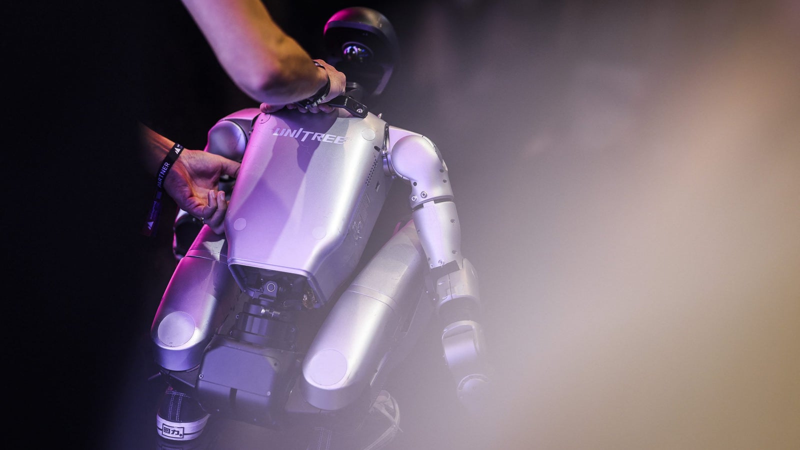 A person adjusting a humanoid robot with a silver body and black features, surrounded by a dim lighting atmosphere.