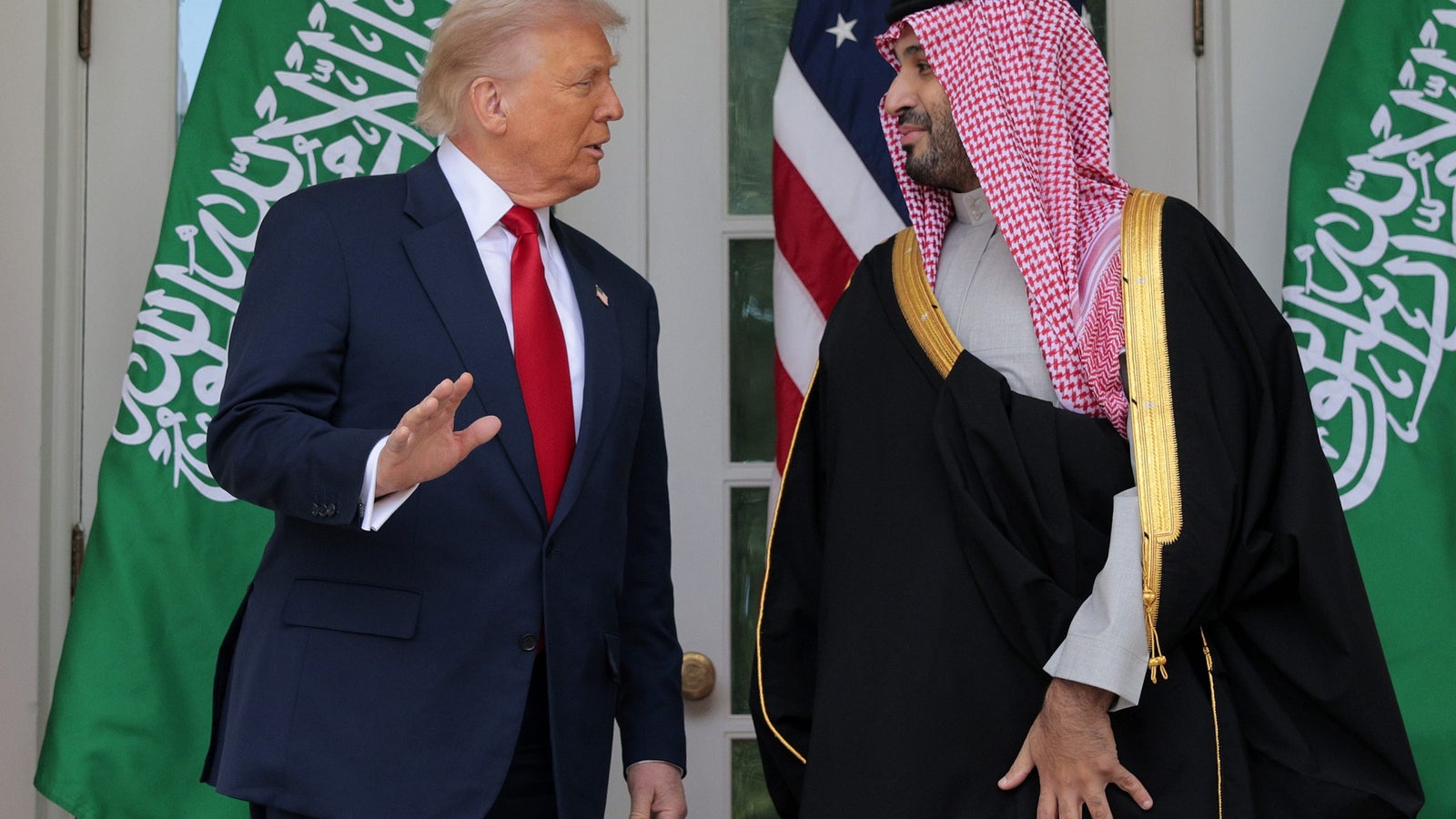 Two men engaging in conversation, with Saudi and American flags in the background.