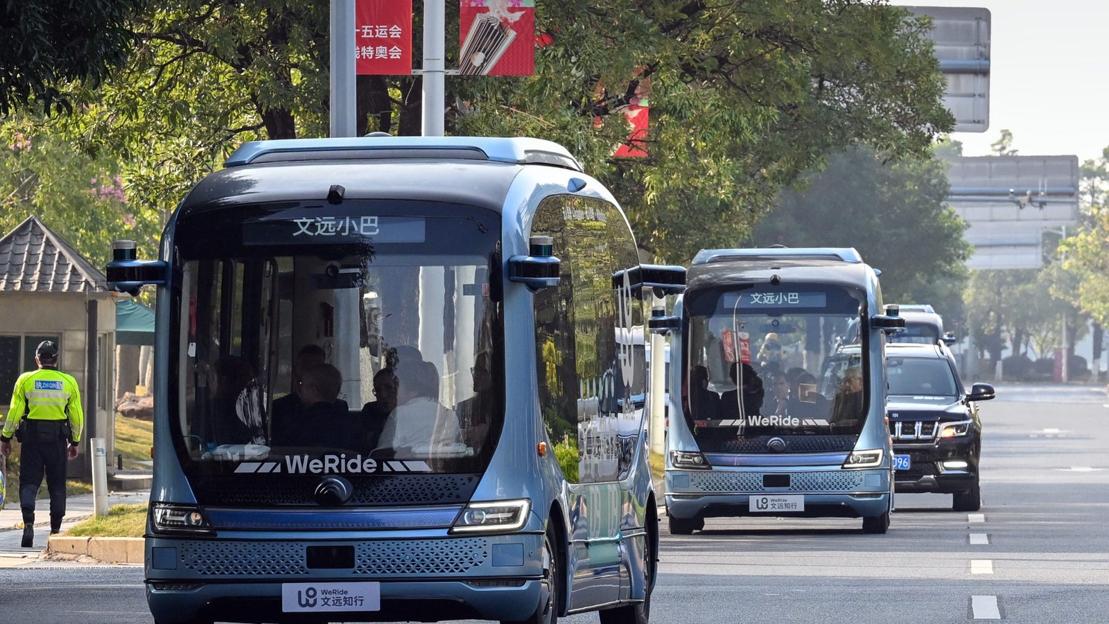 Two autonomous buses are driving down a city street, with trees and buildings visible in the background.