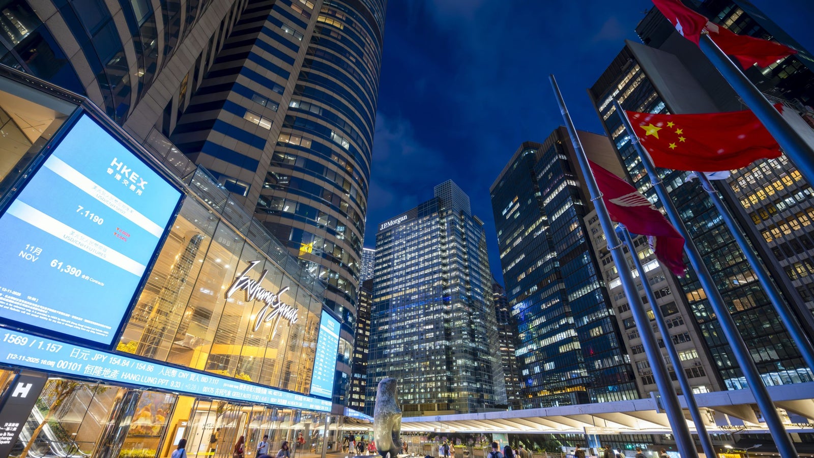 Skyscrapers and bright lights dominate a cityscape at night, featuring a fountain and Chinese flags in the foreground.