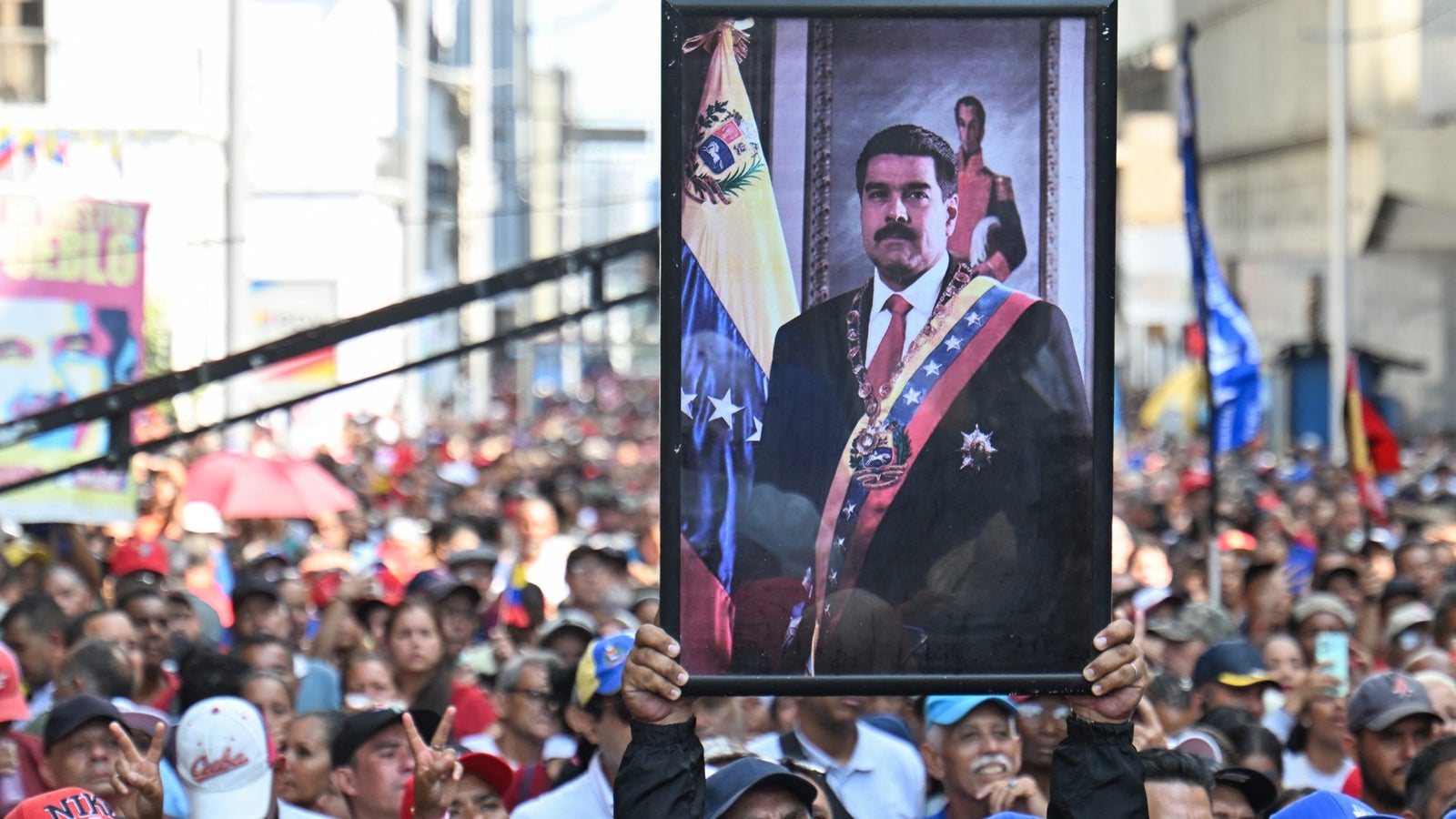 A person holds a framed portrait of a leader amidst a large crowd, with Venezuelan flags and banners in the background.