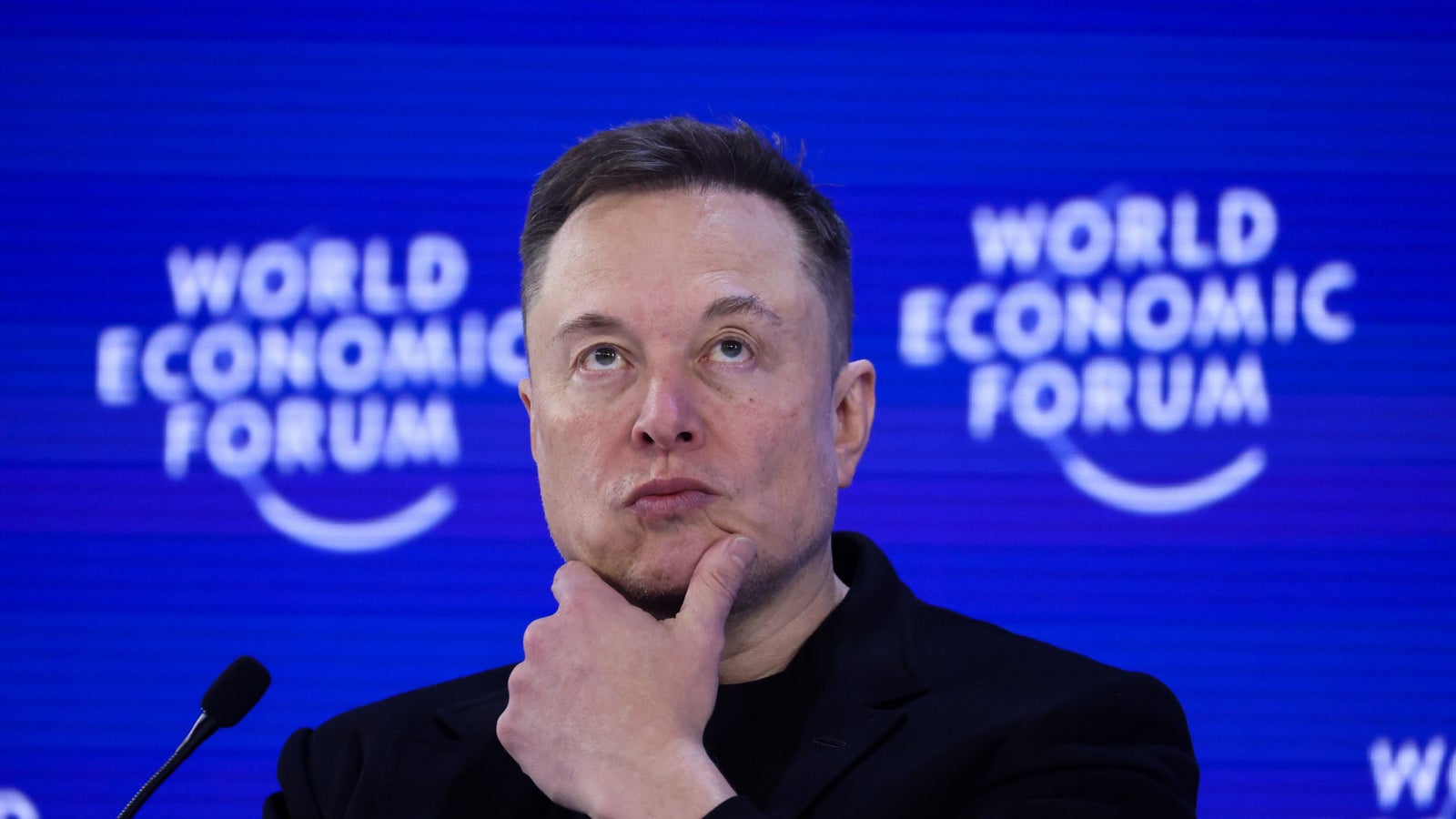 A thoughtful person with short hair is pondering while seated at a podium, with a backdrop reading "World Economic Forum."