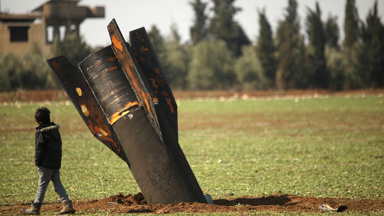 A child walks past a large, partially buried rocket in a green, open field with trees in the background.