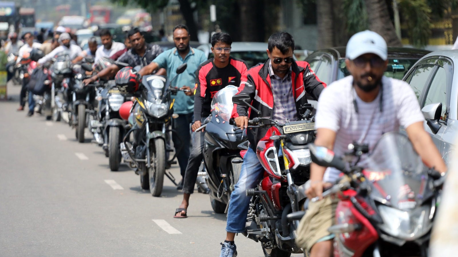Motorcyclists wait in traffic on a busy street, some wearing helmets and casual clothing, with parked cars visible on the side.