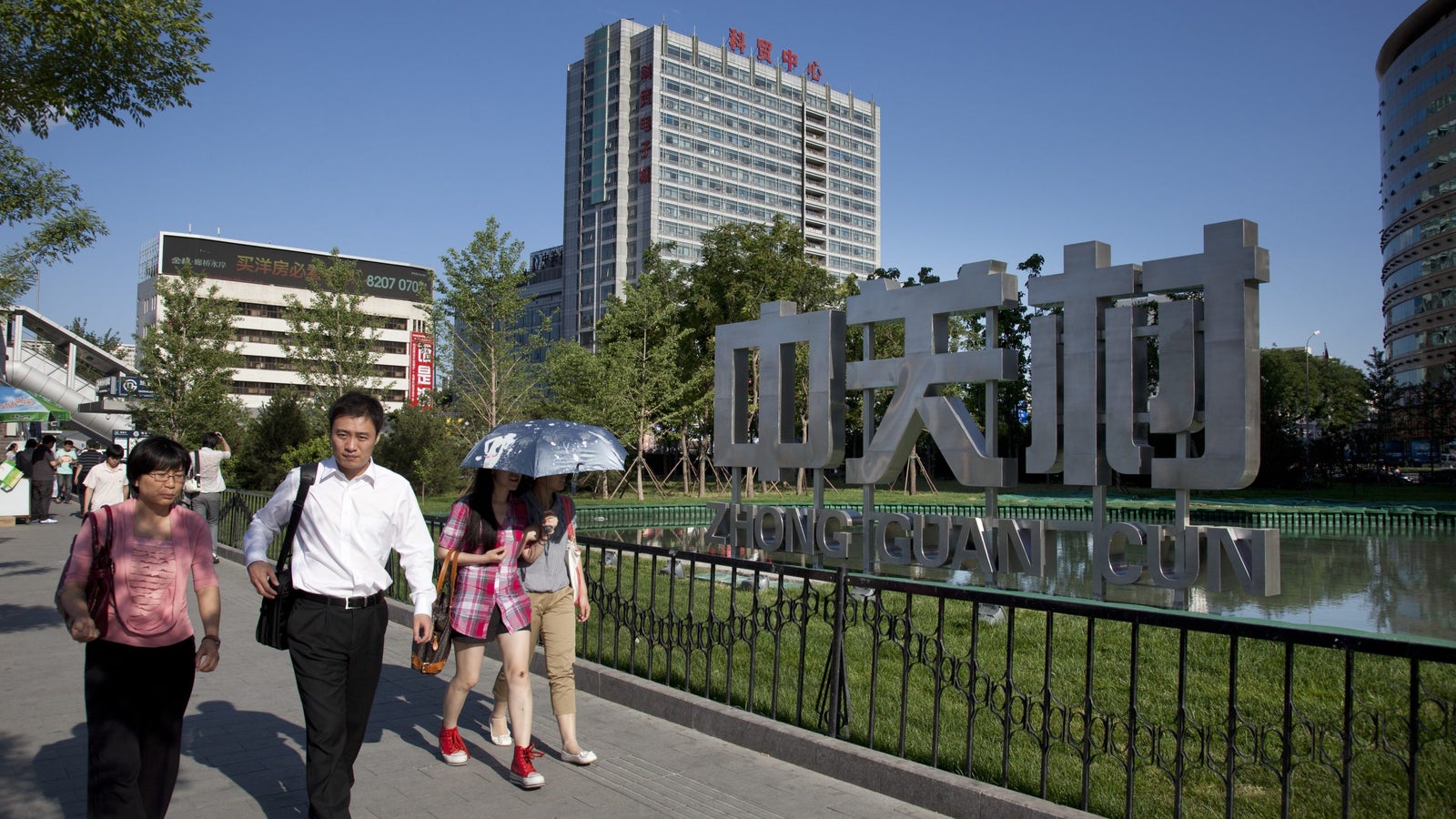 A photo of a man and two women walking on a street, with a tall building and a sign that reads Zhonngguancun in Chinese characters, in the background.