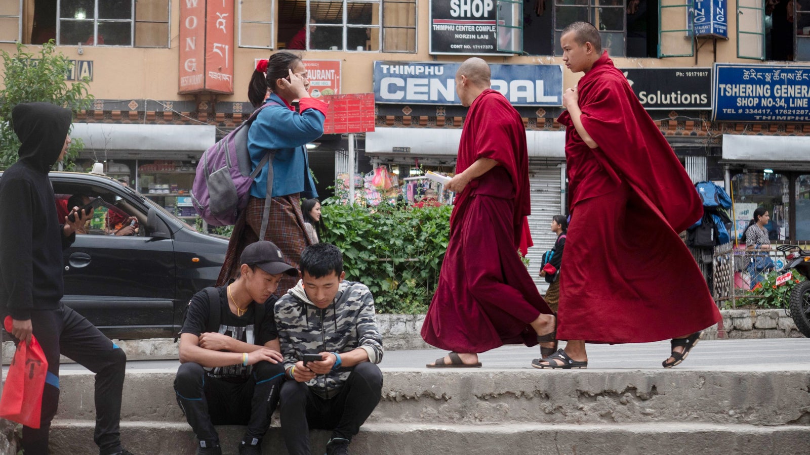 Two monks in red robes walk past a group of young people sitting on steps, with city shops in the background.