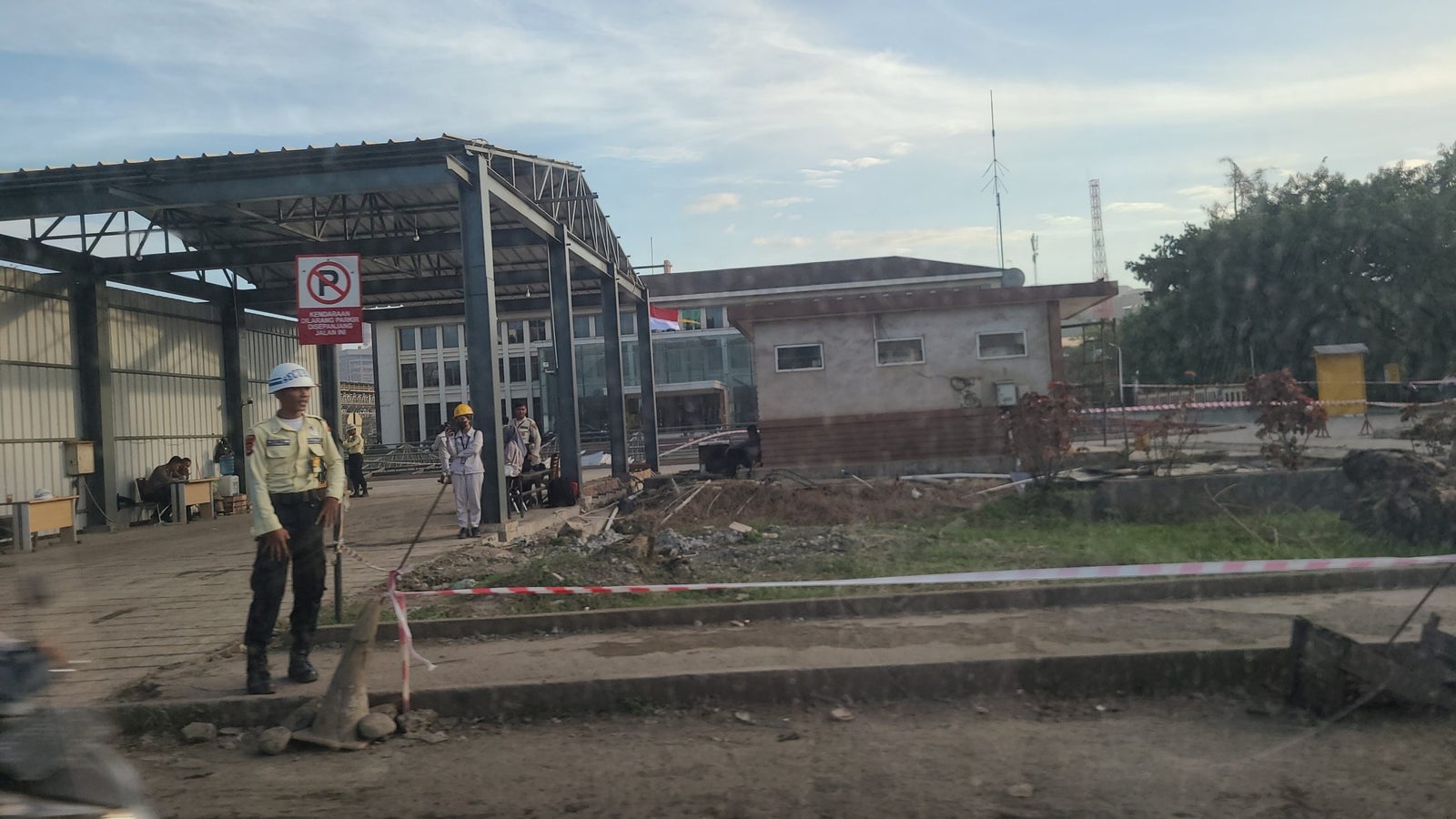 A construction site viewed from a distance, featuring two workers—one in a safety helmet and uniform standing near an empty metal framework and another in white attire and a yellow helmet, all set against a backdrop of a partly built structure and a flag post with flags visible. There is a no parking sign in the background, and construction debris is scattered across the ground.