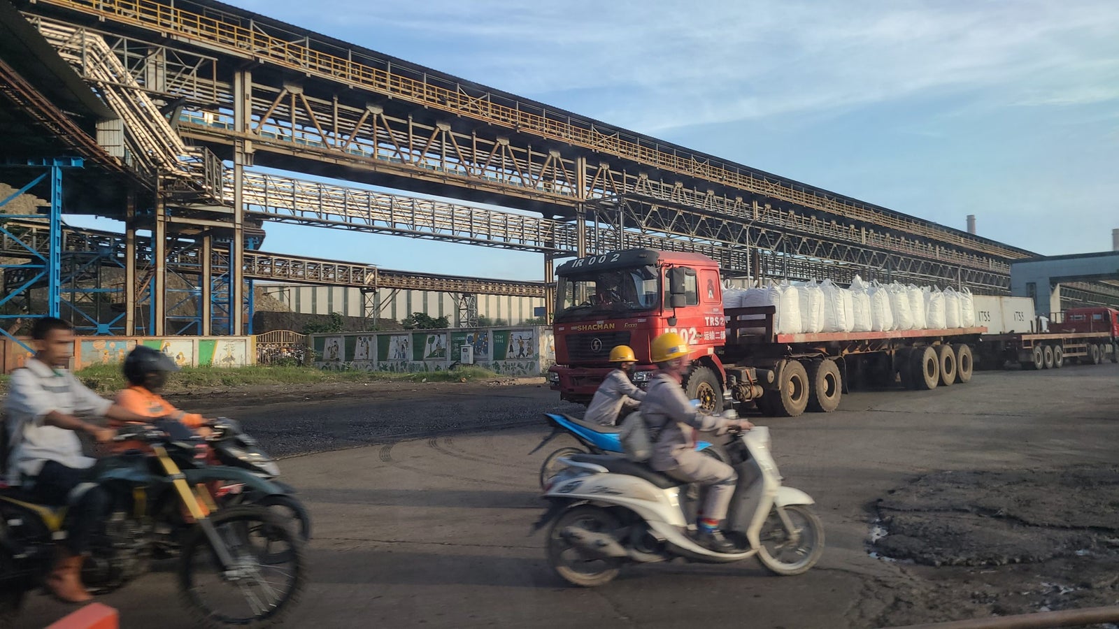A busy industrial area showing a red truck loaded with large white bags parked on a road, while two motorcyclists pass by. There are large structures overhead, likely conveyors, and a background of warehouses and concrete barriers with faded artwork.