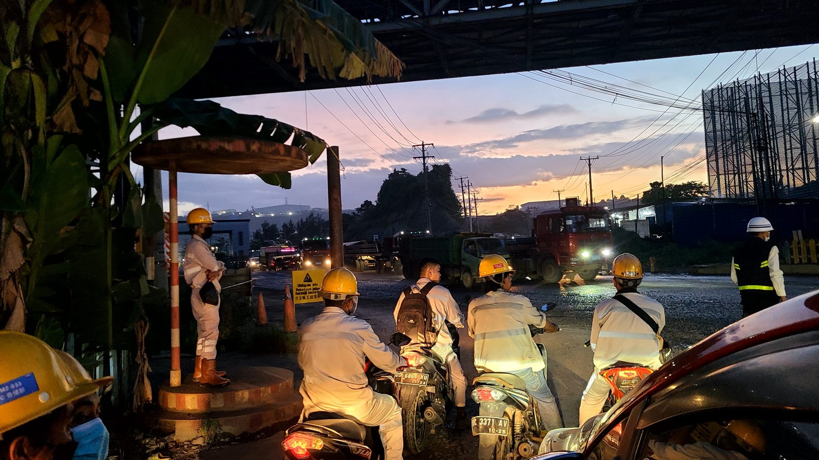 A group of workers in reflective clothing and helmets are waiting on motorcycles at a construction site during twilight, with power lines and vehicles in the background. The scene is illuminated by the fading light of the sunset.