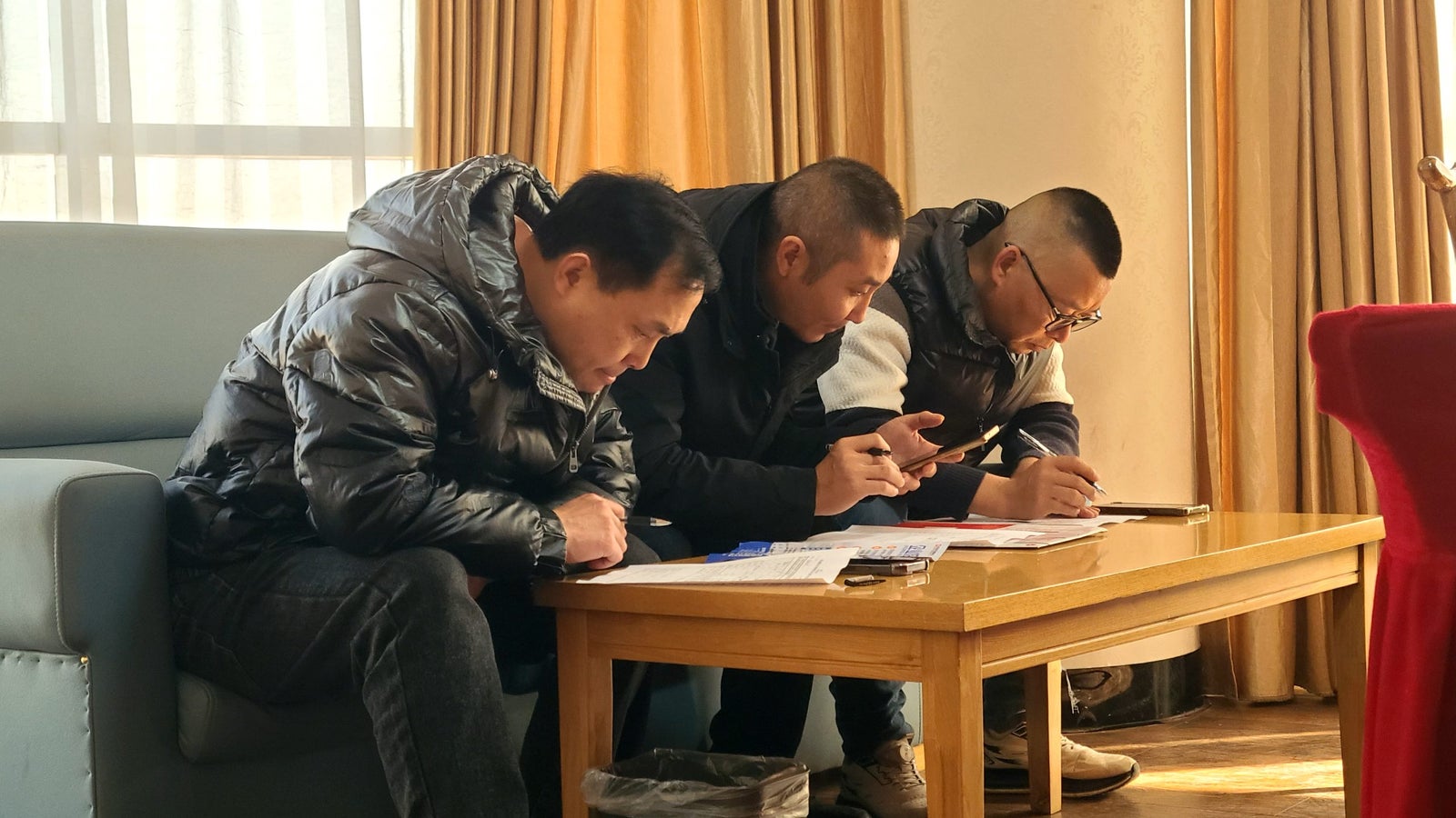 Three men seated on a sofa, focused on their phones and writing on a table in a well-lit room with curtains and wooden flooring.