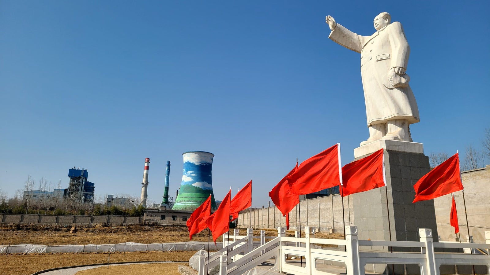 A large white statue of a figure with one arm raised stands on a pedestal, surrounded by several red flags. In the background, there are industrial structures including cooling towers painted with clouds and smokestacks. The sky is clear and blue.