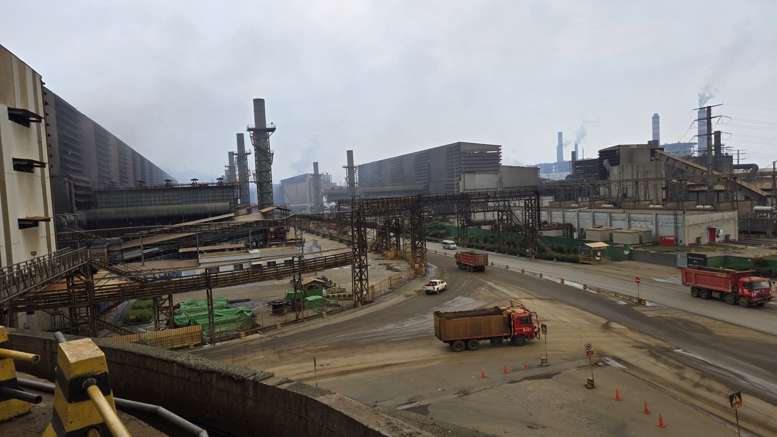 A view of an industrial facility with tall smokestacks and extensive metal structures, alongside dirt roads and construction materials, under a cloudy sky. Trucks are visible on the roads, along with traffic cones and signs.