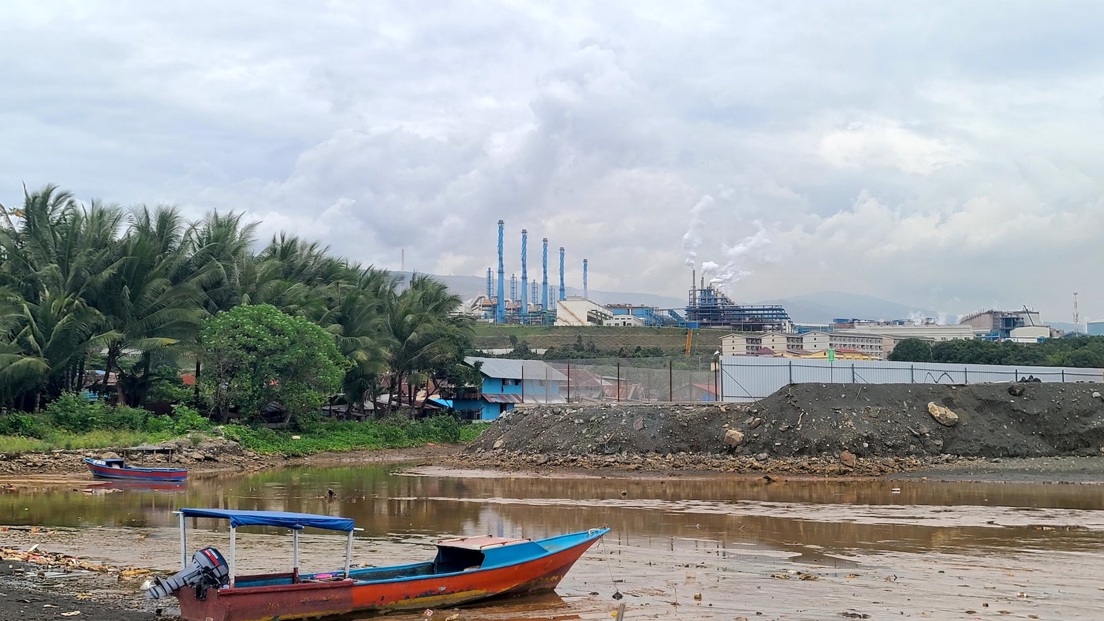 A view of a muddy river with two colorful fishing boats in the foreground and a backdrop of industrial buildings and smokestacks under a cloudy sky.