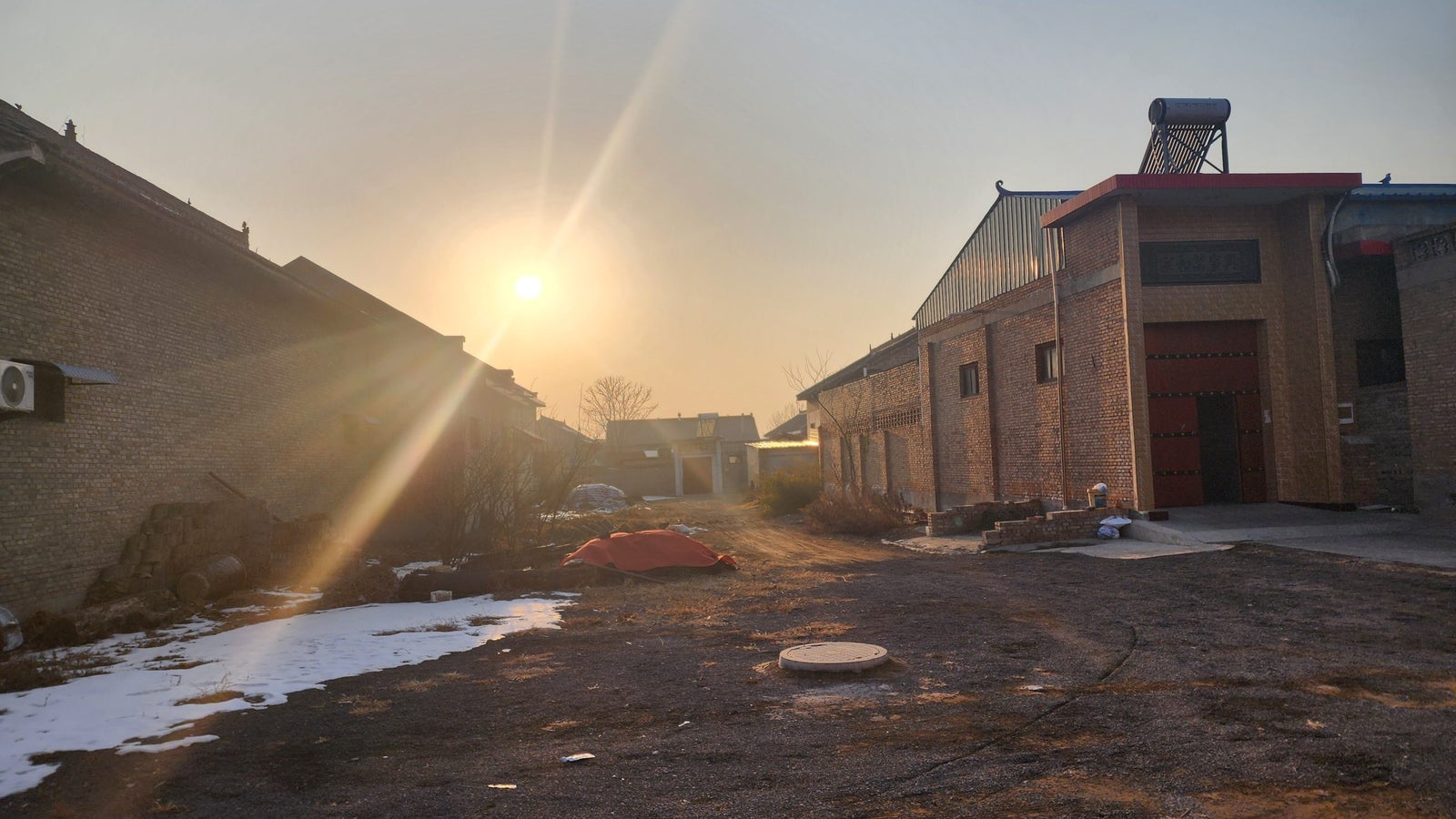 A sun sets behind buildings in an industrial area, casting rays of light across the scene, with a mix of snow and dirt on the ground and an orange tarp in the foreground.