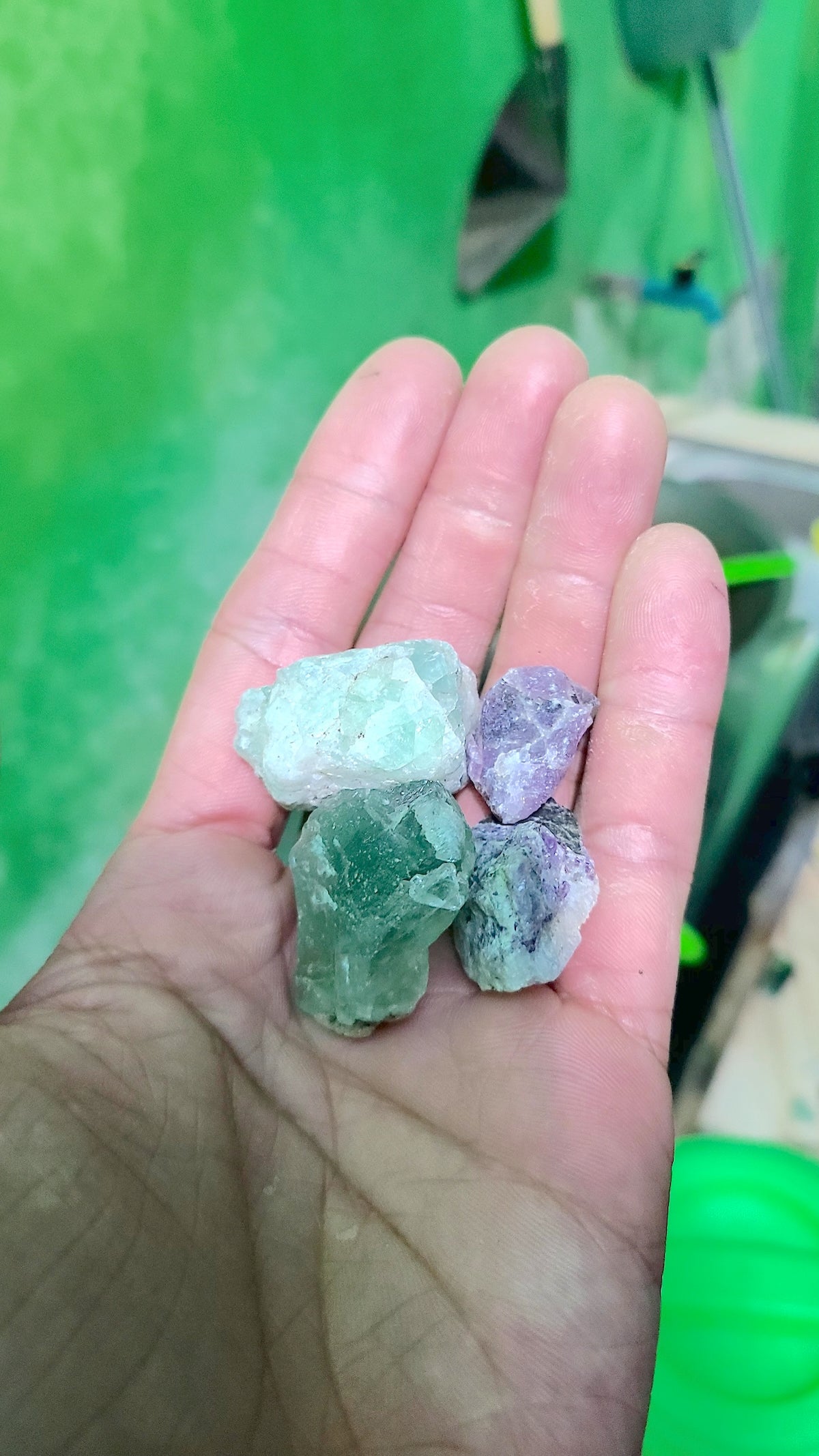 A hand holding several colorful crystals, including green and purple, against a bright green background with tools and a green container partially visible.