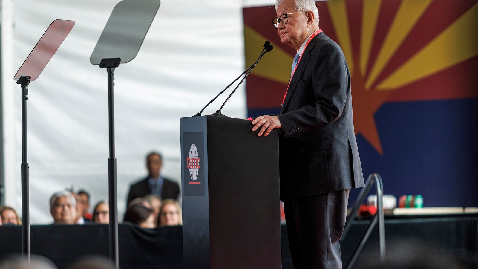 A photograph showing a man wearing a suit, standing at a podium with the TSMC logo.