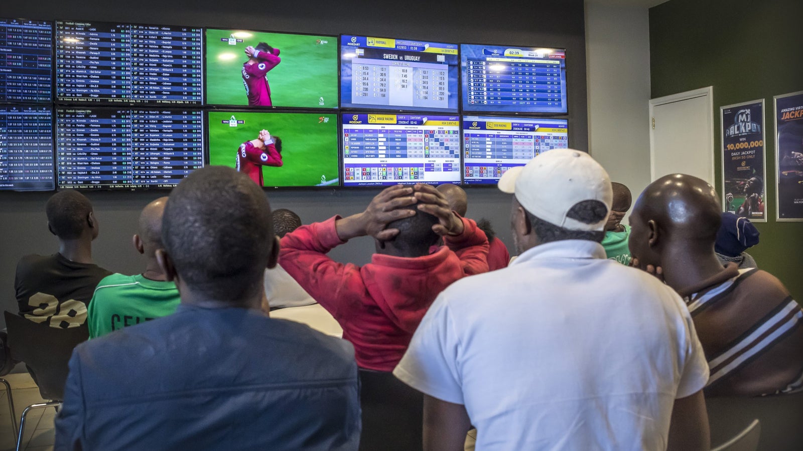 A group of people in a 'Betting office' intently watch a football match and betting screens.