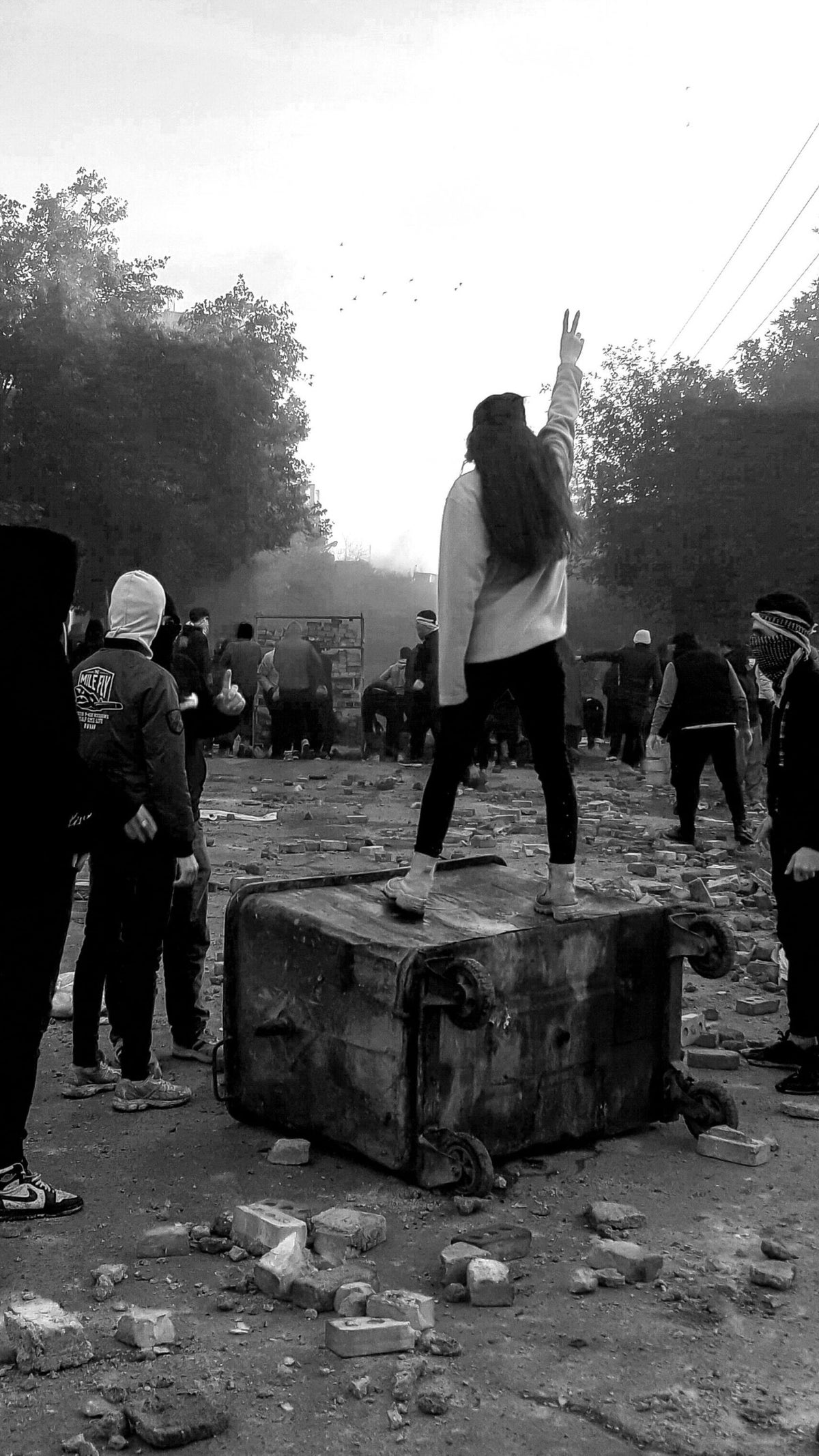 A black and white photograph showing a personal standing atop a large trash bin, holding up two fingers in a 'v' alongside a number of other people during a protest.