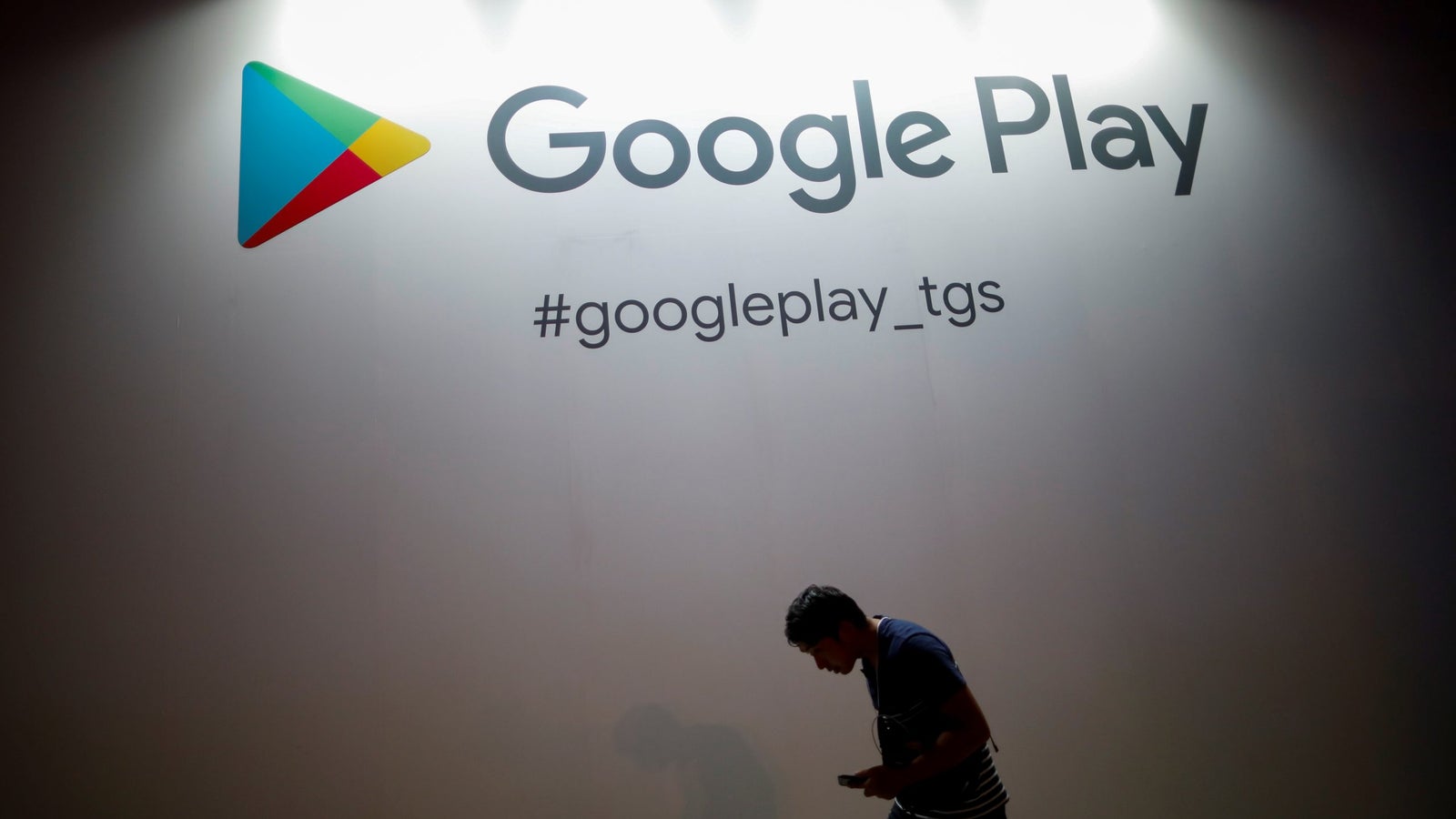 The logo of Google Play lit up on a wall with the silhouette of a person walking past it.