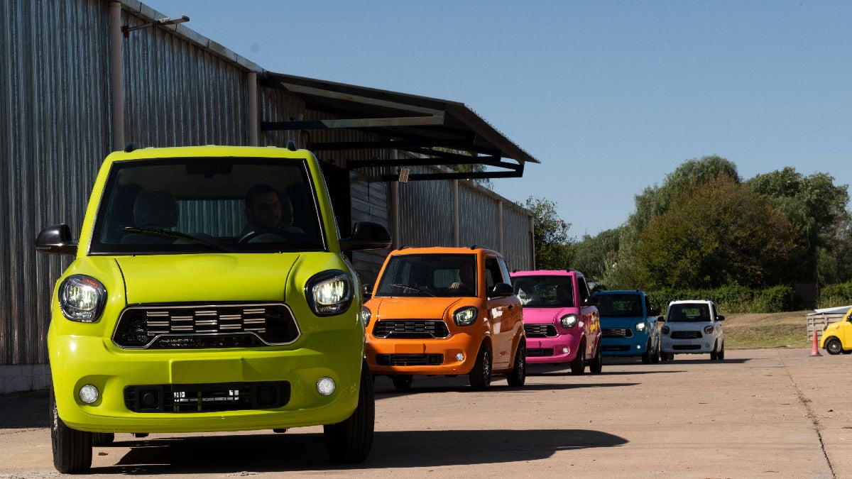 A line of colorful compact cars parked on a concrete surface, featuring a bright green car in the foreground, followed by orange, pink, blue, and white cars. The scene is set outside a metal building with trees in the background under a clear blue sky.