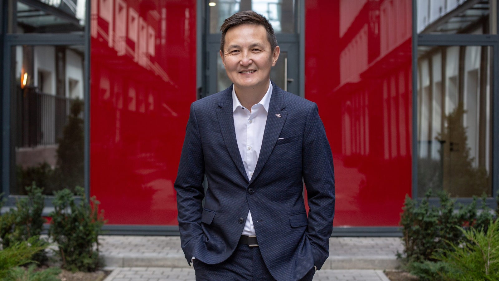 A man in a dark suit stands with his hands in his pockets in front of a bright red wall, smiling at the camera. The setting features modern architecture with greenery in the foreground.