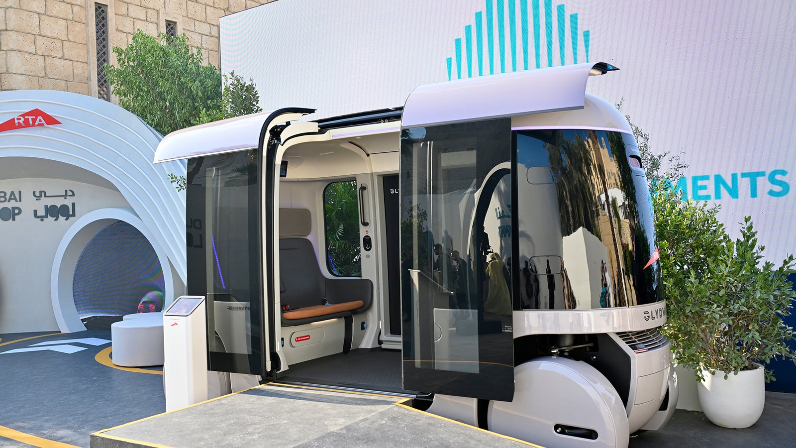 A futuristic autonomous vehicle with sleek design, open doors, and a modern interior, displayed at an event venue.