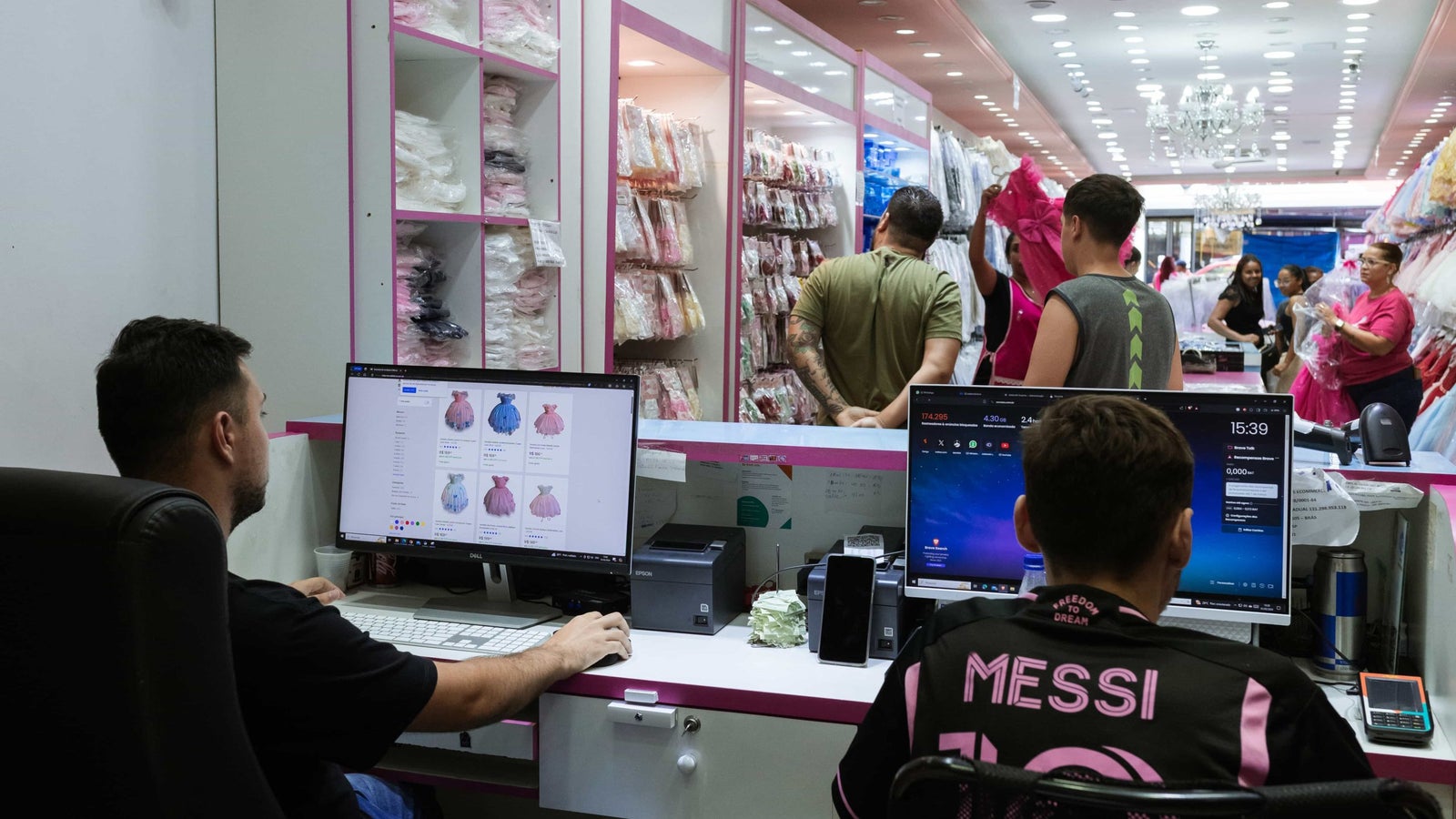 Two clothing store worker sits behind the register using computers, one of the screens shows an ecommerce store page.