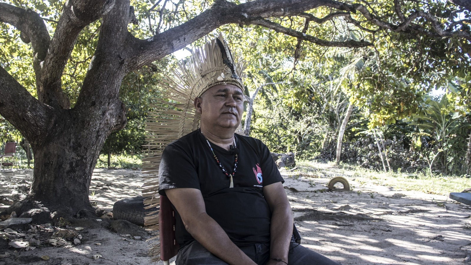 A man wearing a traditional feathered headdress and a black t-shirt sits on a chair under a large tree in a natural outdoor setting. He has a serious expression and is surrounded by greenery and sandy ground.